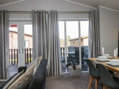 A living room with grey curtains a black sofa a dining table set with white dishes and an outdoor patio with wicker furniture at Lakeside 20 in Warton near Carnforth