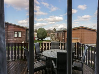 A balcony with a round table and six chairs overlooking wooden lodges and a small lake at Lakeside 20 in Warton near Carnforth