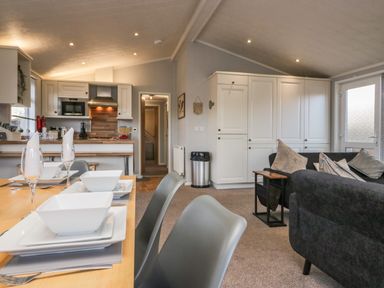 An open-plan living area with a dining table set with white dishware grey chairs a kitchen with white cabinets and wooden countertops and a seating area with a sofa and cushions at Lakeside 20 in Warton near Carnforth