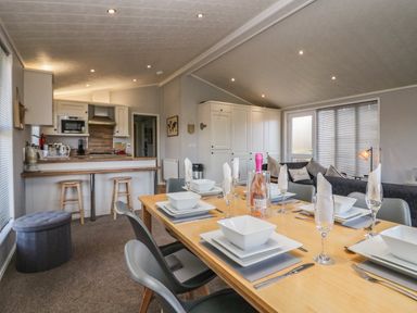 A dining table set with white dishes and glasses next to a kitchen with stools and a living area with a sofa at Lakeside 20 in Warton near Carnforth