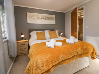 A bedroom with a double bed with orange blanket and pillows bedside tables with lamps and a partially open door at Lakeside 20 in Warton near Carnforth