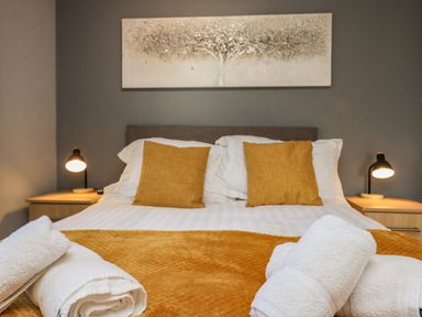A bedroom with a double bed with yellow pillows and blanket a pair of bedside tables with lamps and a painting above the bed at Lakeside 20 in Warton near Carnforth