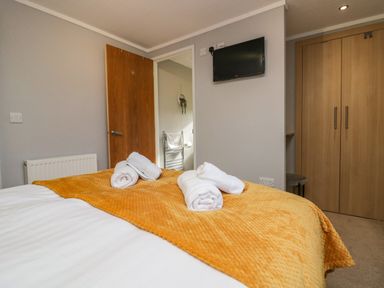 A bedroom with a bed covered by a yellow blanket and rolled white towels on it a wooden door a wall-mounted tv and a wooden wardrobe at Lakeside 20 Warton near Carnforth