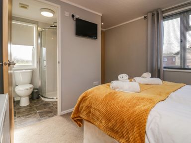 A bedroom with a bed covered in a mustard blanket and rolled towels with an en suite bathroom and a wall-mounted TV at Lakeside 20 in Warton near Carnforth