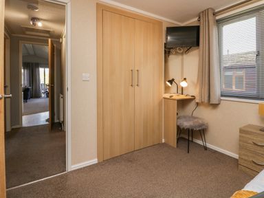 A bedroom with a wooden wardrobe a small desk with two lamps and a furry stool a window with blinds and curtains and a wall-mounted TV at Lakeside 20 in Warton near Carnforth
