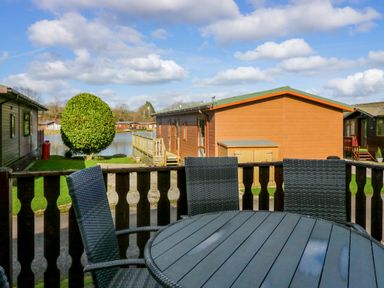 A patio table and chairs on a wooden deck with a view of a lake and wooden lodges at Lakeside 20 in Warton near Carnforth