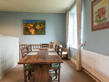 A dining room with a wooden table and chairs next to windows with curtains at Riverbank Cottage in Alnmouth