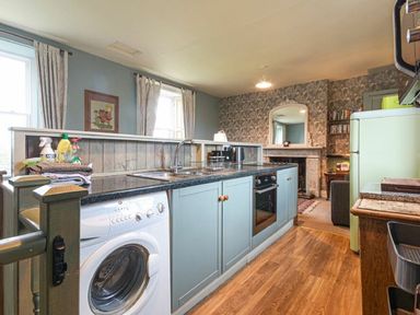 A kitchen area with washing machine oven and sink connected to a living room with fireplace at Riverbank Cottage in Alnmouth