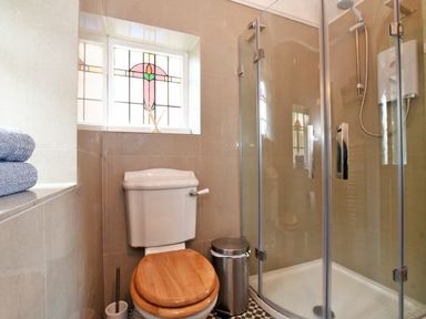 A bathroom with a toilet wooden seat a metal bin glass shower enclosure and stained glass window at Riverbank Cottage in Alnmouth