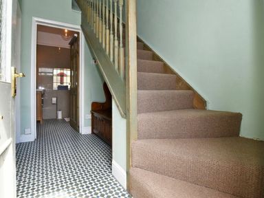 A hallway with stairs on the right and a door leading to a bathroom with a stained glass window at Riverbank Cottage in Alnmouth