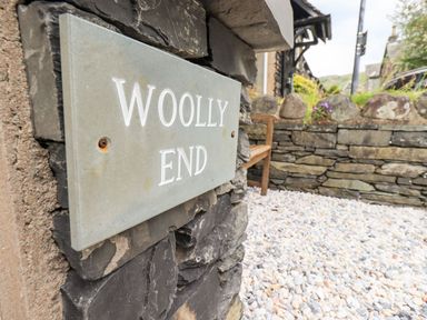 A stone wall with a sign reading Woolly End and a pebble ground area with a wooden bench outside Woolly End Cottage in Ambleside