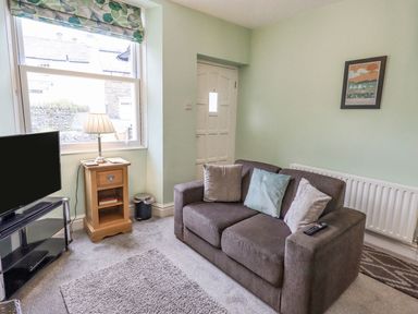 A small living room with a grey sofa and cushions a television on a stand a wooden side table with a lamp and a window at Woolly End Cottage in Ambleside