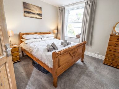 A bedroom with a wooden bed and white bedding with towels on it a window with curtains two bedside tables with lamps and a wooden chest of drawers with a mirror at Woolly End Cottage in Ambleside