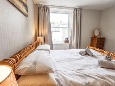 A bedroom with a wooden bed frame white bedding rolled towels a window with curtains and a wooden dresser with a mirror at Woolly End Cottage in Ambleside