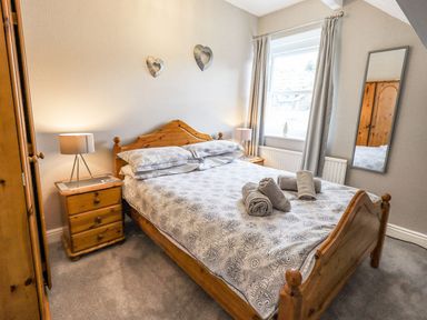 A bedroom with a wooden bed a patterned duvet towels on the bed nightstands with lamps a window with curtains and a wall mirror at Woolly End Cottage Ambleside