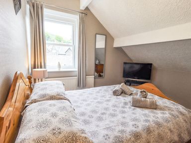 A bedroom with a patterned bedspread a wooden bed frame a window with curtains a lamp a wall mirror and a television at Woolly End Cottage in Ambleside