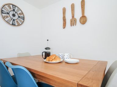 A dining area with a wooden table blue and gray chairs a plate of croissants cups plates a coffee press a wall clock and wooden kitchen utensils on the wall at The Old Forge in Trerulefoot