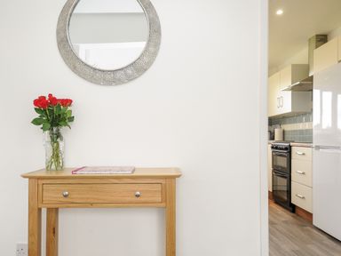 A wooden table with a vase of red flowers and a book a round mirror on the wall and a view into a kitchen with appliances and cupboards at The Old Forge in Trerulefoot