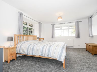 A bedroom with a wooden double bed, two bedside tables with lamps, a wooden chest, large windows with curtains, and a grey carpet at The Old Forge in Trerulefoot