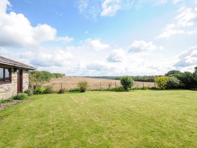 A grass lawn with bushes and a stone building on the left and a field and sky in the background at The Old Forge in Trerulefoot