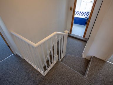 A staircase with gray carpet and white wooden railing leading to a hallway with blue and white tiled bathroom in the background at Ayr Farmhouse in St Ives