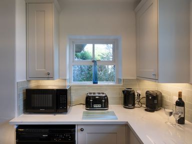 A kitchen counter with a microwave toaster kettle coffee machine and wine bottle with glasses under a window at Ayr Farmhouse in St Ives