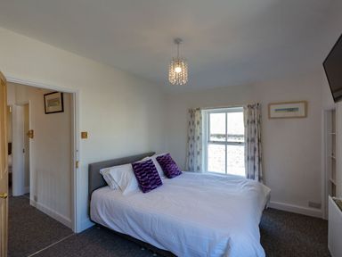 A bedroom with a double bed and purple cushions a window with patterned curtains and a wall-mounted tv at Ayr Farmhouse in St Ives