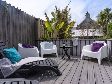 An outdoor deck with white chairs and black lounge chairs with colorful pillows and a small black table with wine glasses at Ayr Farmhouse in St Ives