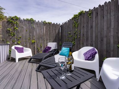 An outdoor seating area with white chairs and black lounge chairs with cushions and a table with wine glasses and bottle at Ayr Farmhouse in St Ives