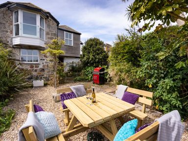 An outdoor patio area with wooden benches and a table with cushions and wine glasses near a red barbecue and a stone house at Ayr Farmhouse in St Ives