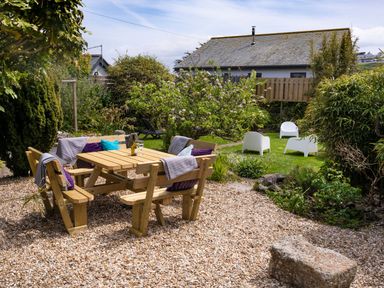 An outdoor garden area with a wooden picnic table and benches with cushions and throws surrounded by gravel and green lawn with white chairs and plants at Ayr Farmhouse in St Ives