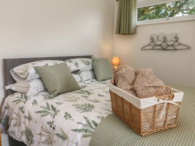 A bedroom with a bed covered with leafy patterned bedding a wicker basket with blankets bedside lamp and window at Jasmine Bydown near Barnstaple