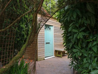 A wooden building with a light blue door beside a wooden bench visible through a metal arch among green plants at Jasmine Bydown near Barnstaple