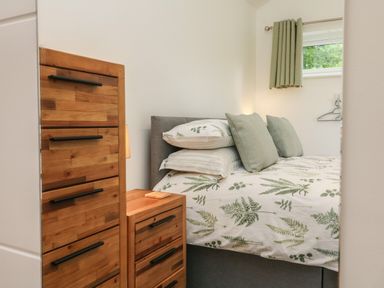 A bedroom with a bed featuring leafy patterned bedding next to wooden dressers and a small window with green curtains at Fern Bydown near Barnstaple