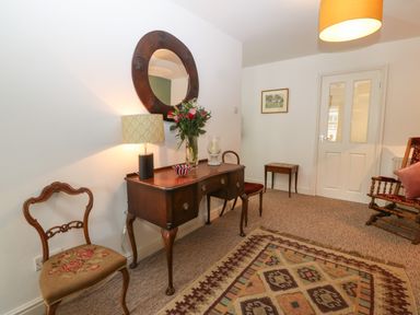 A room with wooden chairs a wooden table with a lamp flowers and a bowl a round mirror on the wall and a patterned rug on the floor at The Bell Apartment in Cemaes Bay