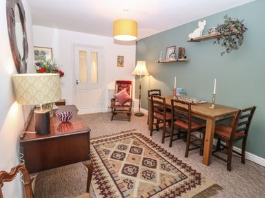A dining room with a wooden table and chairs a patterned rug and a sideboard with a lamp and flowers at The Bell Apartment in Cemaes Bay