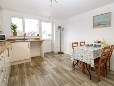 A kitchen with wooden chairs around a table with a leaf patterned tablecloth and a countertop with a plant and microwave at The Bell Apartment in Cemaes Bay