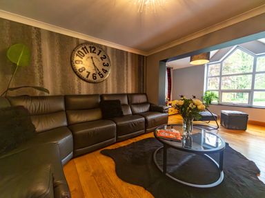 A living room with a leather sectional sofa a round glass coffee table with flowers and magazines a large wall clock and a window with a view at Parc Y Deri Rhos-y-garth near Aberystwyth