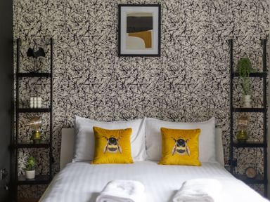 A bedroom with a bed with bee pillows against a wall with abstract wallpaper and shelves holding plants and lamps at Parc Y Deri in Rhos-y-garth near Aberystwyth