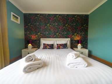 A bedroom with a double bed with white linens and towels on it two bedside tables with lamps and a floral wallpaper at Parc Y Deri in Rhos-y-garth near Aberystwyth
