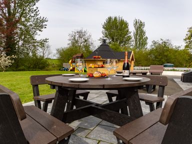An outdoor picnic table set with empty wine glasses, plates, pastries, and a bottle near a wooden hut with trees and playground equipment in the background at Parc Y Deri Rhos-y-garth near Aberystwyth