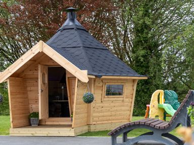 A wooden cabin with a black shingled roof and an open door with flower pots outside and a plastic children's slide and a wooden bench nearby at Parc Y Deri in Rhos-y-garth near Aberystwyth