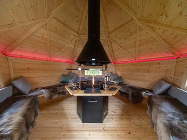 An interior of a wooden cabin with a central black stove a kettle benches with fur covers and pillows at Parc Y Deri in Rhos-y-garth near Aberystwyth
