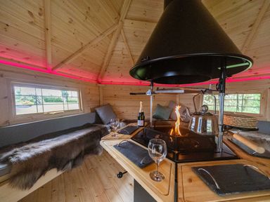 An interior of a wooden cabin with a central fireplace surrounded by wooden benches and cushions at Parc Y Deri in Rhos-y-garth near Aberystwyth