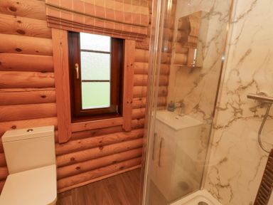 A bathroom with wooden log walls a window a toilet a shower with glass door and a white sink with marble wall panels at 24 Lakeside Drive in Felton