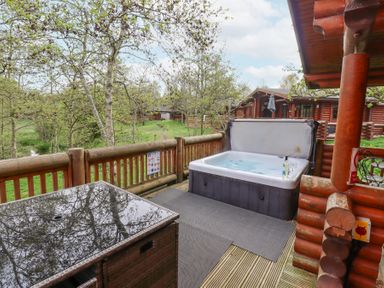 An outdoor deck with a hot tub a table and chairs overlooking trees and wooden cabins at 24 Lakeside Drive in Felton