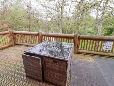 A wooden deck with a glass top table and woven chairs surrounded by a wooden railing and trees at 24 Lakeside Drive in Felton
