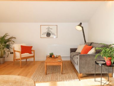 A living room with a gray sofa and armchair with orange cushions a wooden coffee table plants and a floor lamp at Church Farm Dairy Cadbury near Thorverton