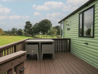 An outdoor wooden deck with a table and two chairs next to a green building with trees and grass in the background at Swan in Shobdon