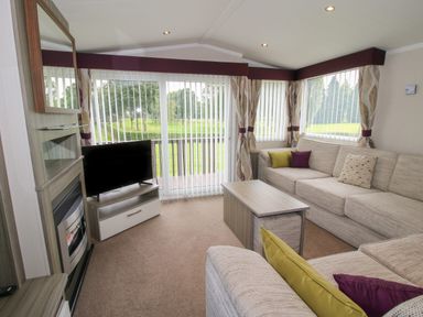 A living room with a beige sectional sofa, coffee table, TV on a stand, fireplace, and large windows with vertical blinds at Swan in Shobdon